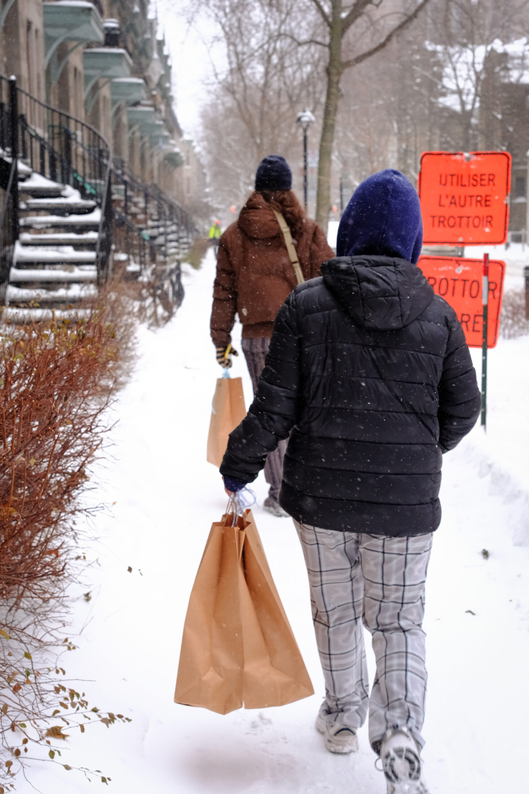 Two volunteers walk down a snowy Montreal sidewalk carrying grocery bags.