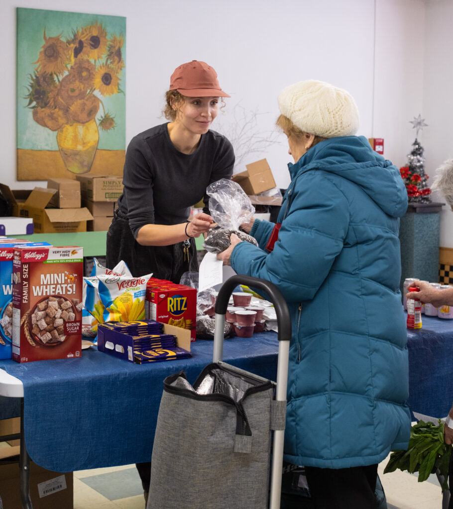 A woman hands a senior a bag of beans, across a table with pantry staples like crackers, cereal, chocolate and apple sauce.