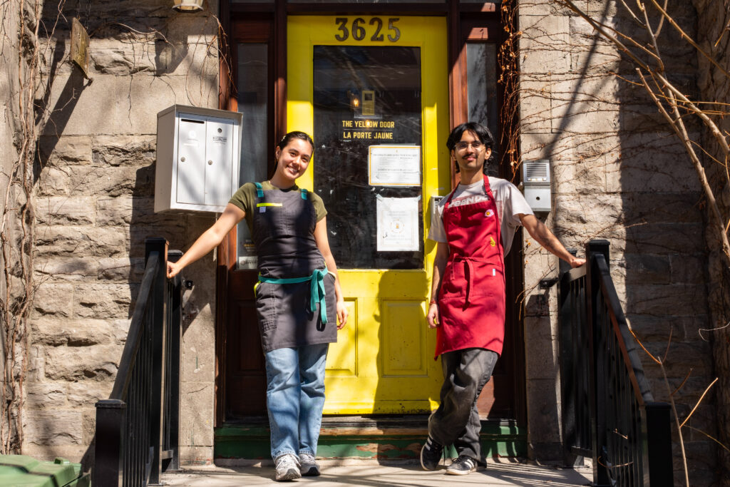 Two Rabbit Hole volunteers stand on the steps of The Yellow Door wearing aprons.
