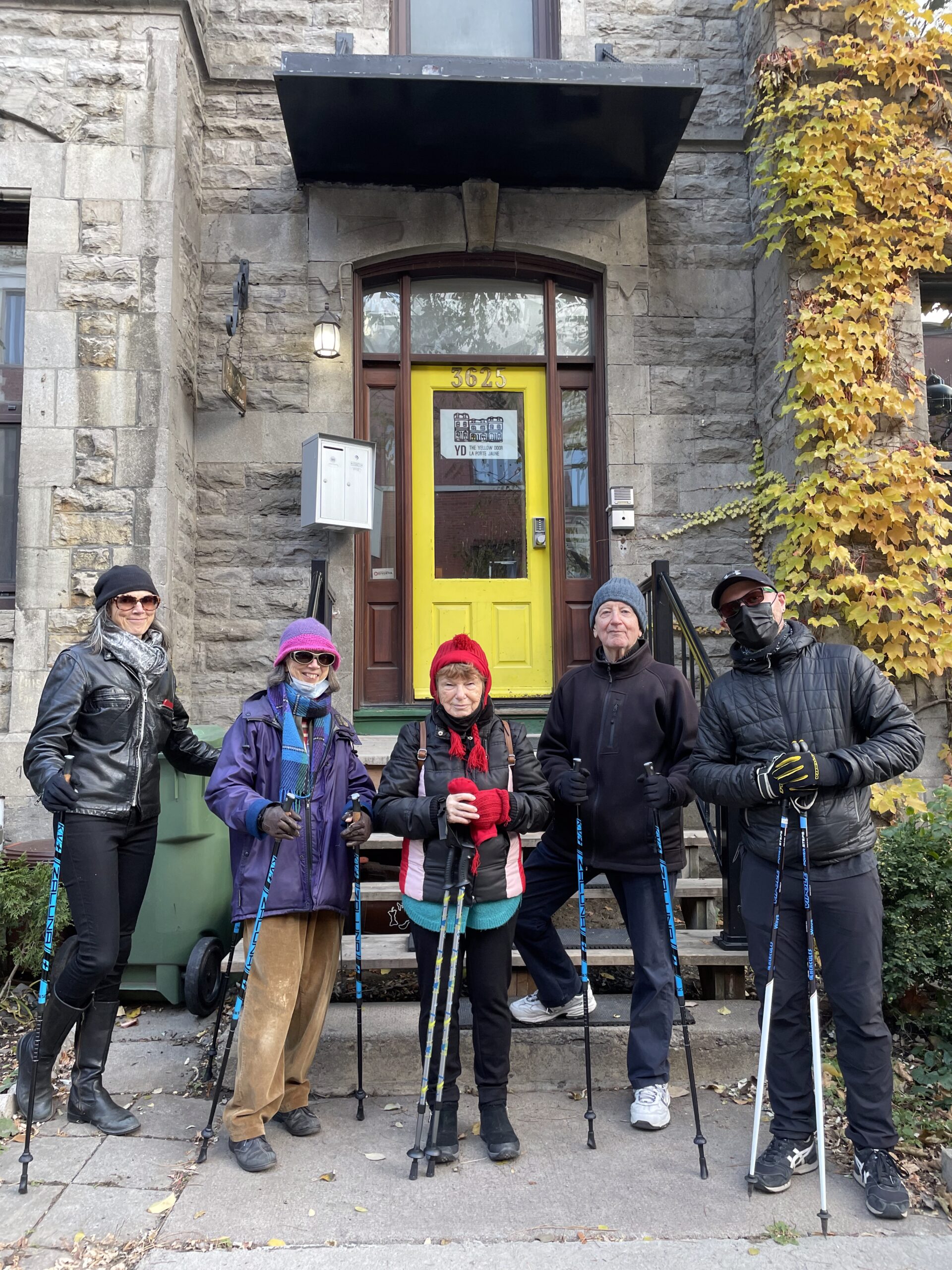 A group of 5 older adults are standing in front of The Yellow Door holding nordic walking poles.